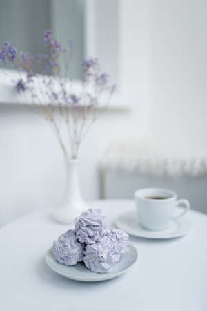 Lilac marshmallow lies on a plate with a cup of coffee and a vase on a white table.