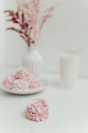 Pink homemade marshmallow lies on a plate on a white background with a white vase and a glass of...
