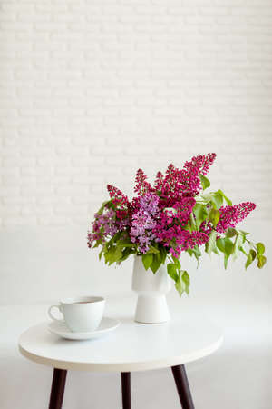 A white cup of tea stands on a white table, next to a bouquet of purple lilacs. Breakfast in a bright room.の写真素材
