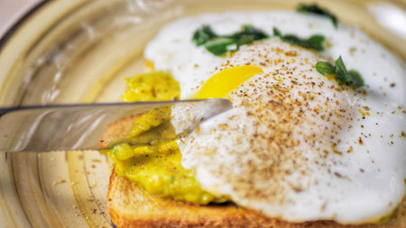Cutting poached egg with runny egg yolk over bread toast with mashed avocado spread