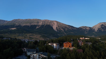 As dusk settles, a charming village rests peacefully against the backdrop of towering mountains, bathed in warm golden light. Nature and architecture blend harmoniously.の写真素材