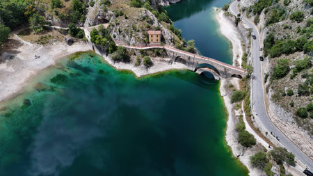 This captivating landscape showcases an antique bridge arching gracefully over clear turquoise waters, adjacent to a quaint structure, with scenic hills in the backdrop.の写真素材