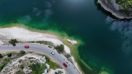 A picturesque drive unfolds alongside a clear turquoise lake, framed by verdant hills and rocky outcrops, radiating tranquility under a sunny sky.の写真素材