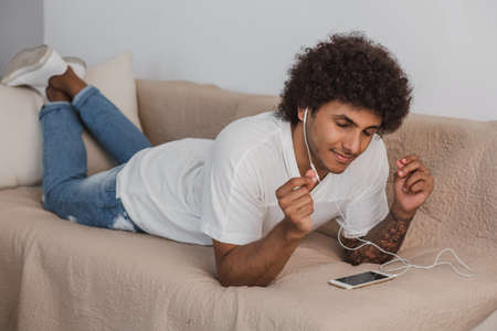 Curly haired stylish man eyes closed lying on the couch at home, enjoy the music.の写真素材