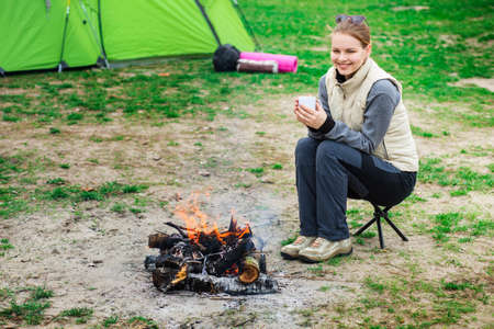 Smiling woman dressed in trekking clothes warm up near the campfire holding a hot cup of tea. Active outdoor recreation, holiday camping.の写真素材