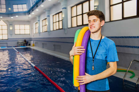 Fit smiling man holding foam roller at swimming pool. Young swimming instructorの写真素材