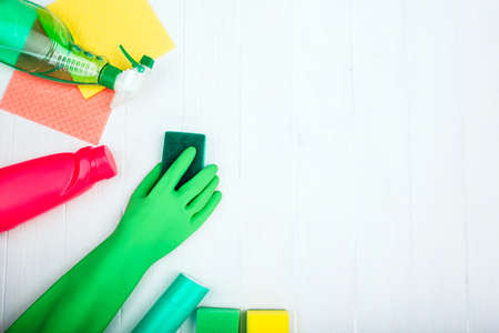 Cleaning products and hand in rubber gloves on white wooden background with empty space, top view