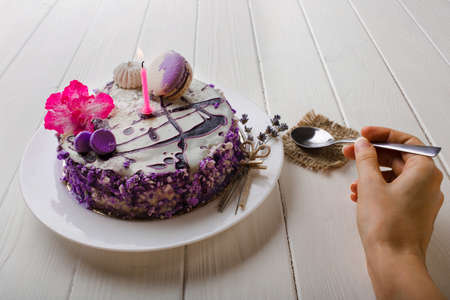 close-up, female hand holding a spoon near the birthday cake with a candle on white wooden table background. Top viewの写真素材