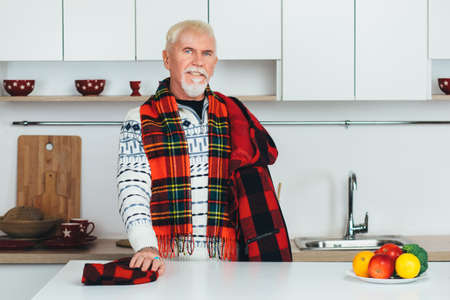 elderly man standing in outerwear, checkered scarf at home in the kitchen, looking at cameraの写真素材