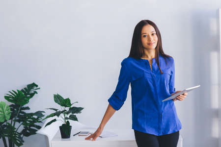 Modern, confident young woman in stylish clothes holding digital tablet, smiling while standing near her working place at office.の写真素材
