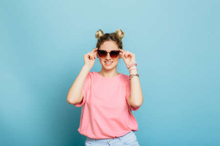 pretty young woman wearing pink clothes adjusting her sunglasses and smiling, while standing against blue background. Looking just perfect.の写真素材