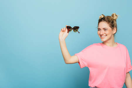 pretty young woman wearing pink clothes holding her sunglasses, smiling and looking away, while standing against blue background. Summer style with copy spaceの写真素材