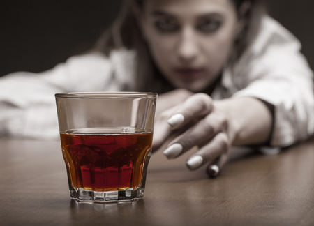 Young woman in depression, drinking alcohol on dark background. Focus on the glassの写真素材