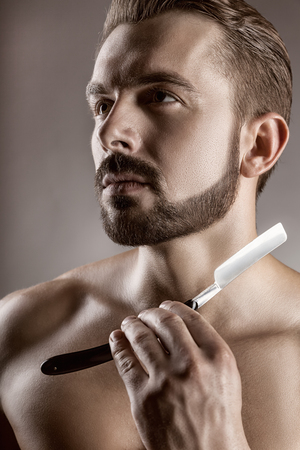 Closeup of young handsome man guy with beard showing razor blade, studio shot. Shave conceptの写真素材