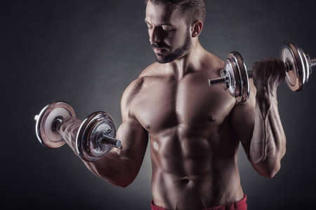 Handsome smiling muscular man exercising with dumbbell on dark backgroundの写真素材