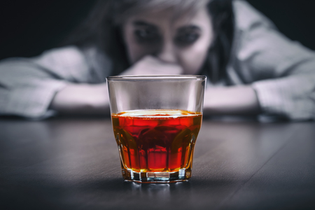 Young woman in depression, drinking alcohol on dark background. Focus on the glassの写真素材