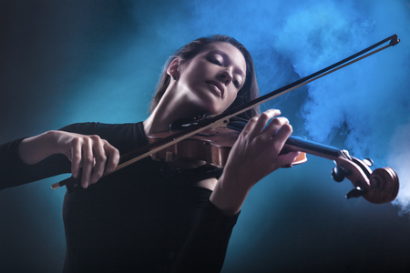 Beautiful young woman playing the violin on dark blue background. Fog in the background. Studio shotの写真素材