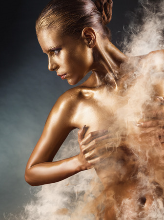 Beautiful gold girl. Fashion woman with golden makeup and bodyart. Fog in the background. Studio shotの写真素材
