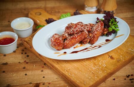 Serving of delicious spicy chicken wings with sesame seeds and sweet chili sauce on white wooden boardの写真素材