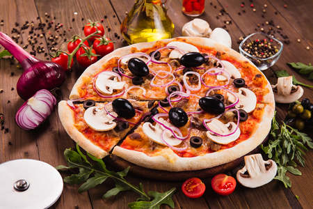 Italian pizza with mushrooms, olives and onion rings with ingredients on wooden table (close). Cut one pieceの写真素材