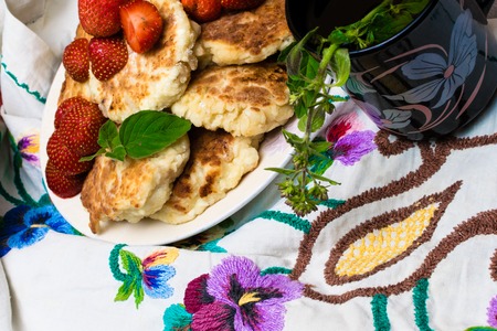 Freshly baked cottage cheese pancakes with raisins in a white plate, cup of tea with mint, melissa twig, embroidered with color flowers tablecloth. Top viewの写真素材