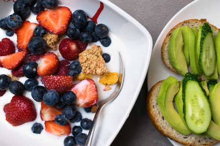 vegetarian breakfast. bowl of oat granola with milk, raw raspberries, blueberries, strawberries, and nuts with spoon for healthy breakfast and avokado and cucumber sandwich top viewの写真素材