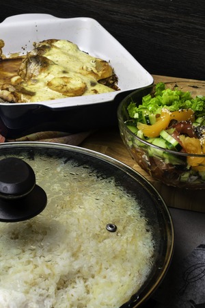 baked raw fish in sour cream, fresh salad with tomatoes, green lettuce, cucumber, sesame seeds, olive oil, rice in pan in wooden boardの写真素材