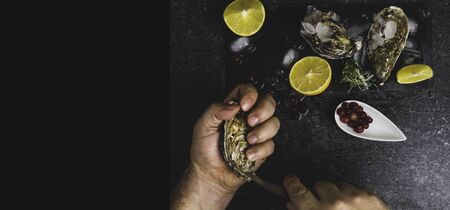 A man with a knife opens a fresh oyster. Dark stone background with lemon, ice, berries, selective focus. Food for men health, rich in protein, iodine, zincの写真素材