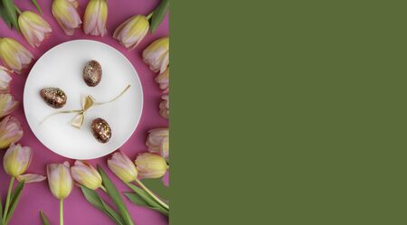 dinner white plate with luxury gold easter eggs, bow, beautiful tulip flowers frame Top view over...