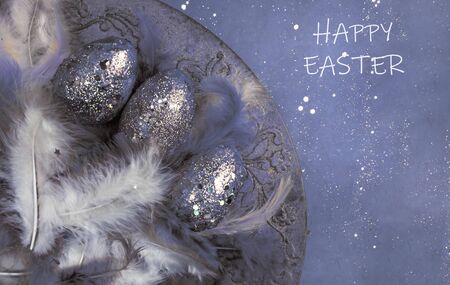 easter eggs, feathers with festive glitter confetti on creative toned in blue color background,...