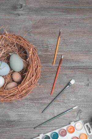 preparing for easter holidays, on wooden table nest with colorful eggs, paints and brushes