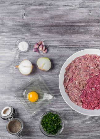 Ingredients for cooking cutlets or burger as raw beef and pork meat on wooden chopping board, onion, dill herbs, salt, pepper, egg, fresh minced meat over woden background, top viewの写真素材