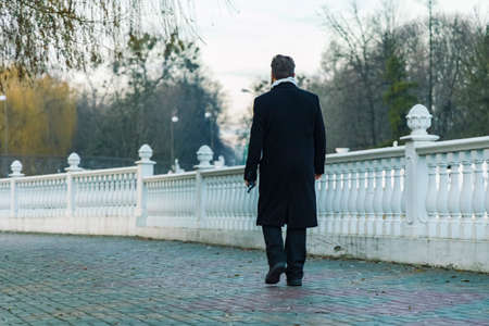 incognito adult man black coat walking park alley paved road depression melancholy mood of early spring time in March with bare trees branchesの写真素材