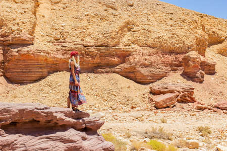 travel photography young woman portrait in long fashion dress stay on edge of cliff in canyon sand stone rocky background environment of Negev Israeli desert dry landscapeの写真素材