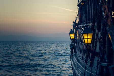 old wooden vintage pirate ship on sea water surface in sunset evening romantic time with yellow light from soft focus lantern in overboard spaceの写真素材