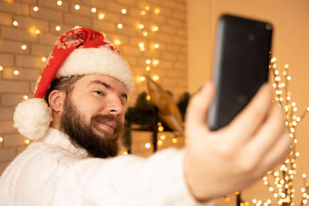 Christmas selfie by middle adult bearded man with white sweater and red hat in warm and cozy decorated room with yellow garland lamps illumination lightingの写真素材