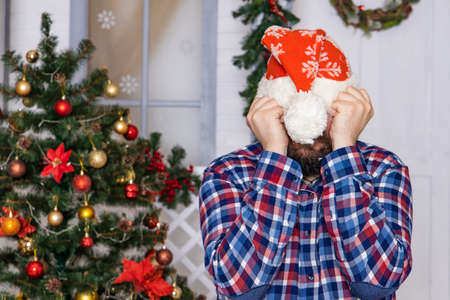 Christmas time emotion of shy guy with hands on hat hiding bearded face festive room background viewの写真素材