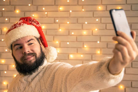 Christmas selfie of bearded man in red hat loneliness in winter holidays concept room interior environment space with garland lights on wall background viewの写真素材