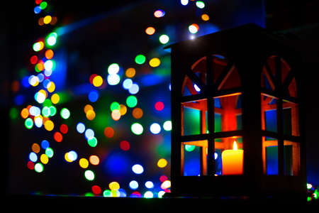 Christmas holidays festive lights inside home in dark room with foreground lantern with candle and background blurred bokeh garland colorful lamps, unfocused concept background pictureの写真素材