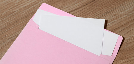 A close-up shot shows a pastel pink envelope slightly open, revealing blank white paper inserts against a textured wooden backgroundの写真素材