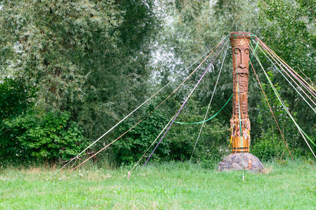 A weathered wooden effigy stands tall in a lush green meadow, adorned with numerous vibrant...