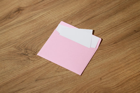 A pastel pink envelope lies open on a textured wooden surface, revealing blank white cards inside,...