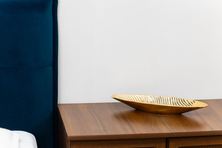A modern bedroom interior featuring a dark wood nightstand with a striking gold leaf-shaped bowl and...