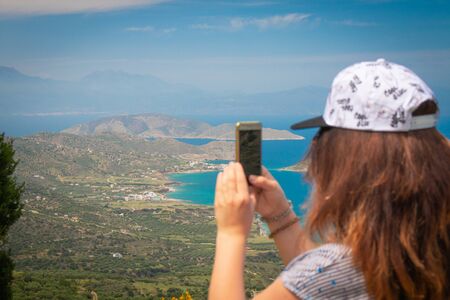 Young woman making photo of a beautiful background of aegean sea and mountains. Woman with brown wavy hair taking picture on vacations.の写真素材