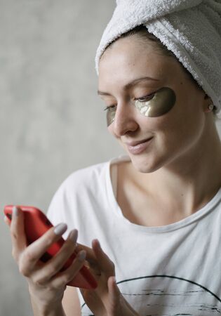 Happy woman with patches under her eyes looks at the phone and smile. Moisturizing and lifting...