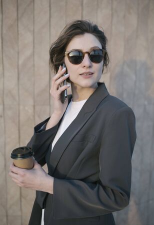 Portrait of a beautiful Caucasian woman in a suit against the urban city background. The girl is talking on the phone and holding a cup of coffee in her handsの写真素材