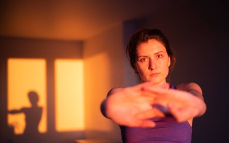 A beautiful girl does stretching exercises at sunset in the home room and looks into the camera. Sunset light, silhouette of a slender girl engaged in sportsの写真素材