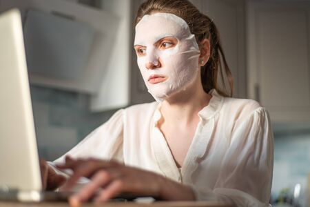 Facial skin care at home during freelance. Young woman typing on a laptop keyboard with a moisturizing mask on her faceの写真素材