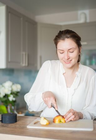 Cheerful woman is cooking in her smart kitchen with a portable speaker and her voice recipe. Cute chef cuts apples to the sounds of the personal audio assistantの写真素材