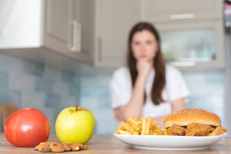 The choice between unhealthy and healthy food. Burger and french fries in front of an apple, tomato and nut. Slender girl in the kitchen trying to make a difficult choice of foodの写真素材